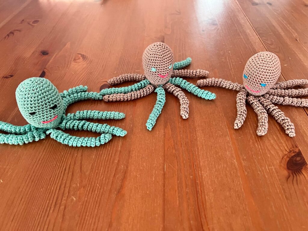Drei gehäkelte Oktopusse in Mint, Beige und Bunt auf einem Holztisch.