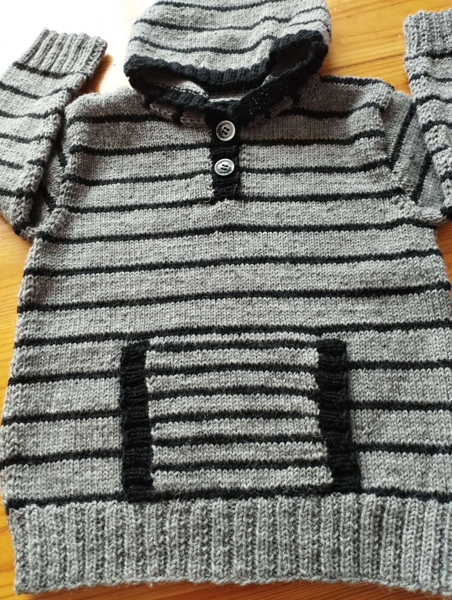 Grauer Strickpullover mit schwarzen Streifen, Kapuze, zwei Knöpfen und einer Bauchtasche auf einem Holztisch.