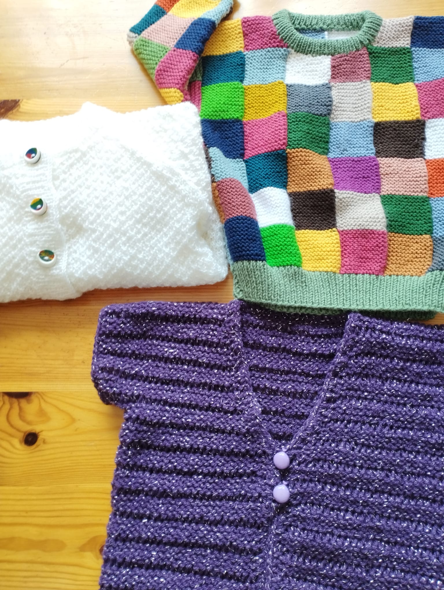 Pullover mit bunten Quadraten neben einer weissen und einer violetten Strickjacke auf einem Holztisch.