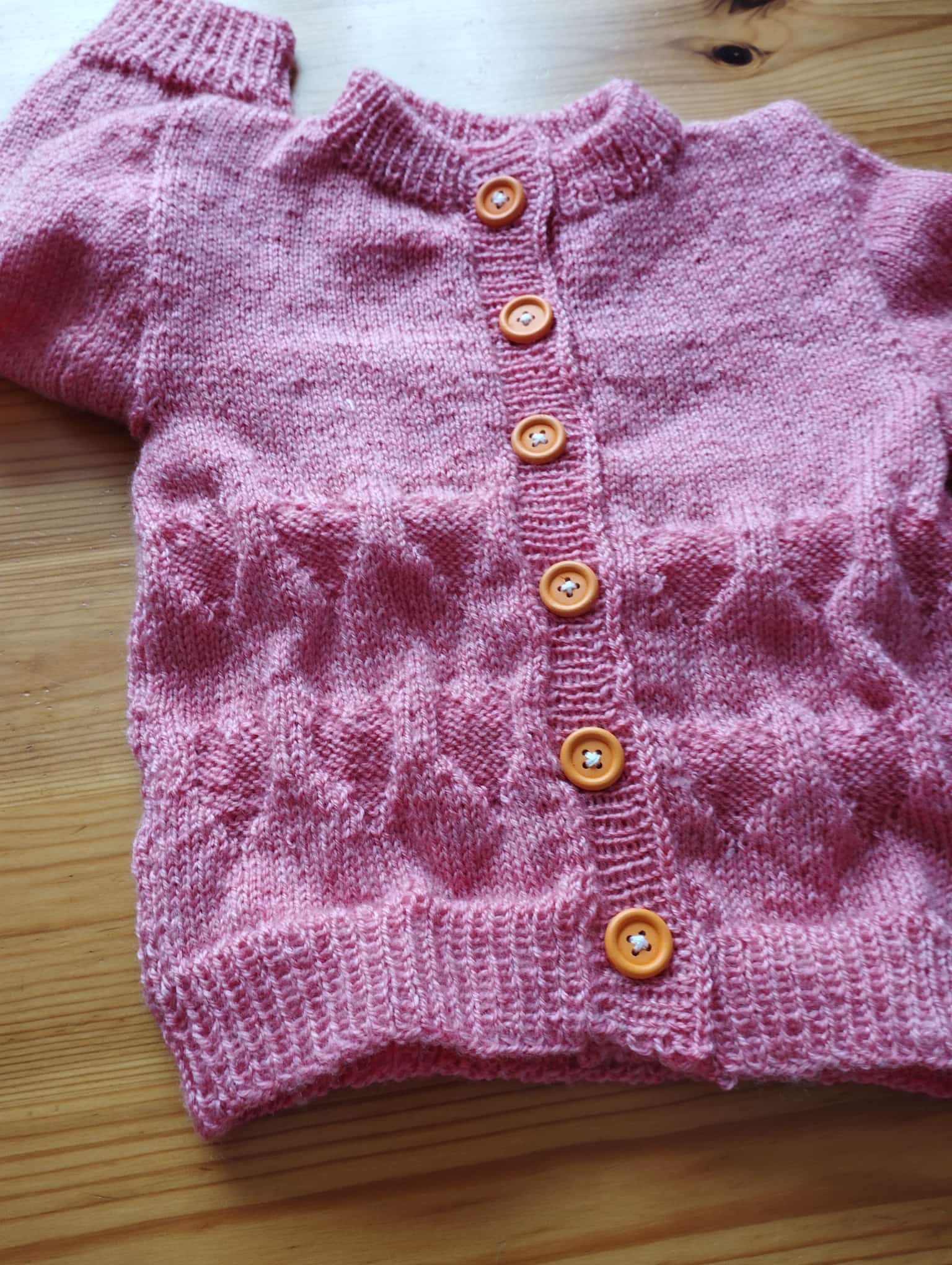 Rosa Strickjacke mit Strukturmuster und sechs orangebraunen Knöpfen auf einem Holztisch.