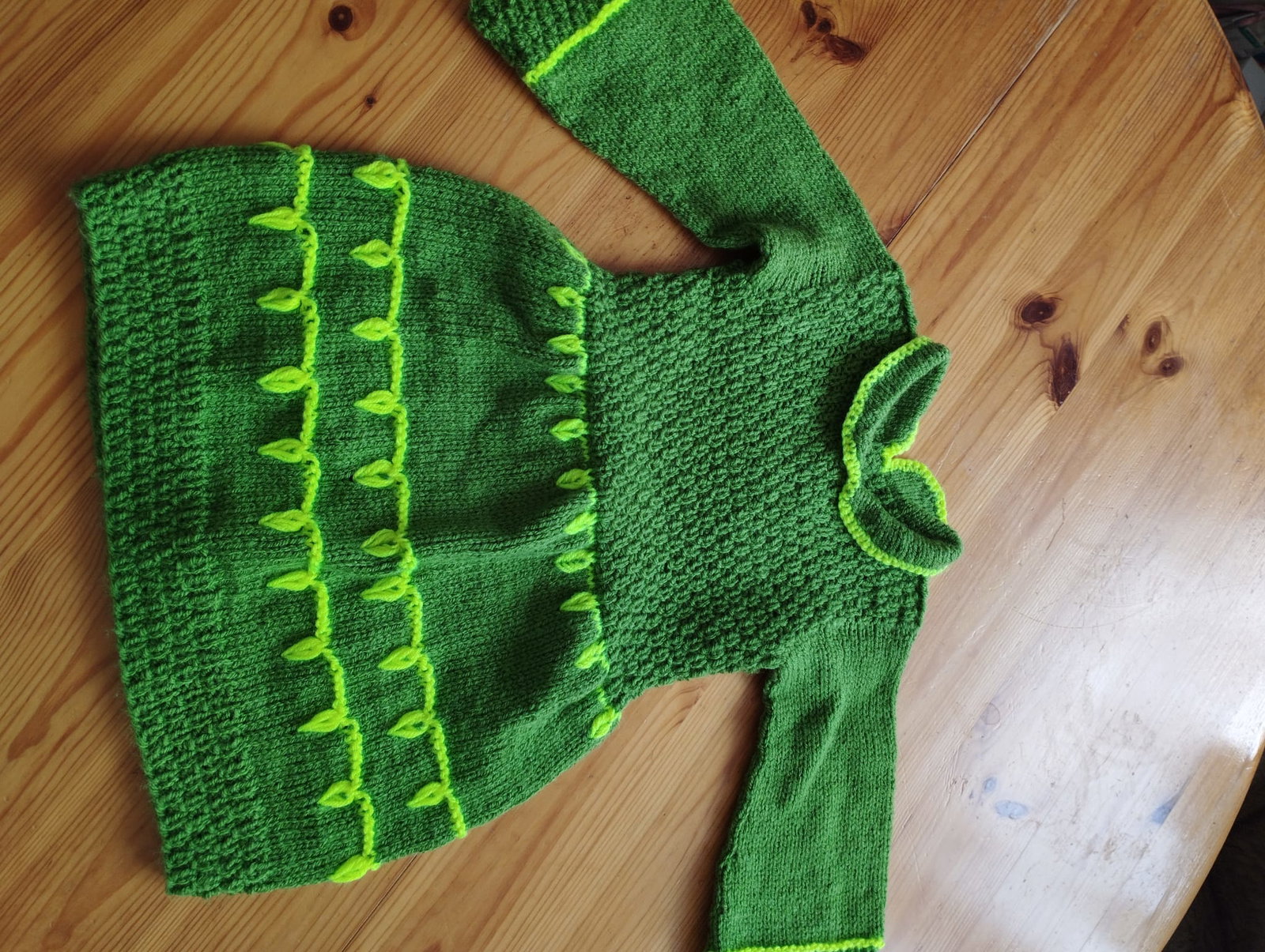 Grünes Strickkleid mit hellgrünem Blattmuster und langen Ärmeln auf einem Holztisch.